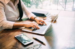 A lady sat writing a blog post at a wooden desk with her phone nearby